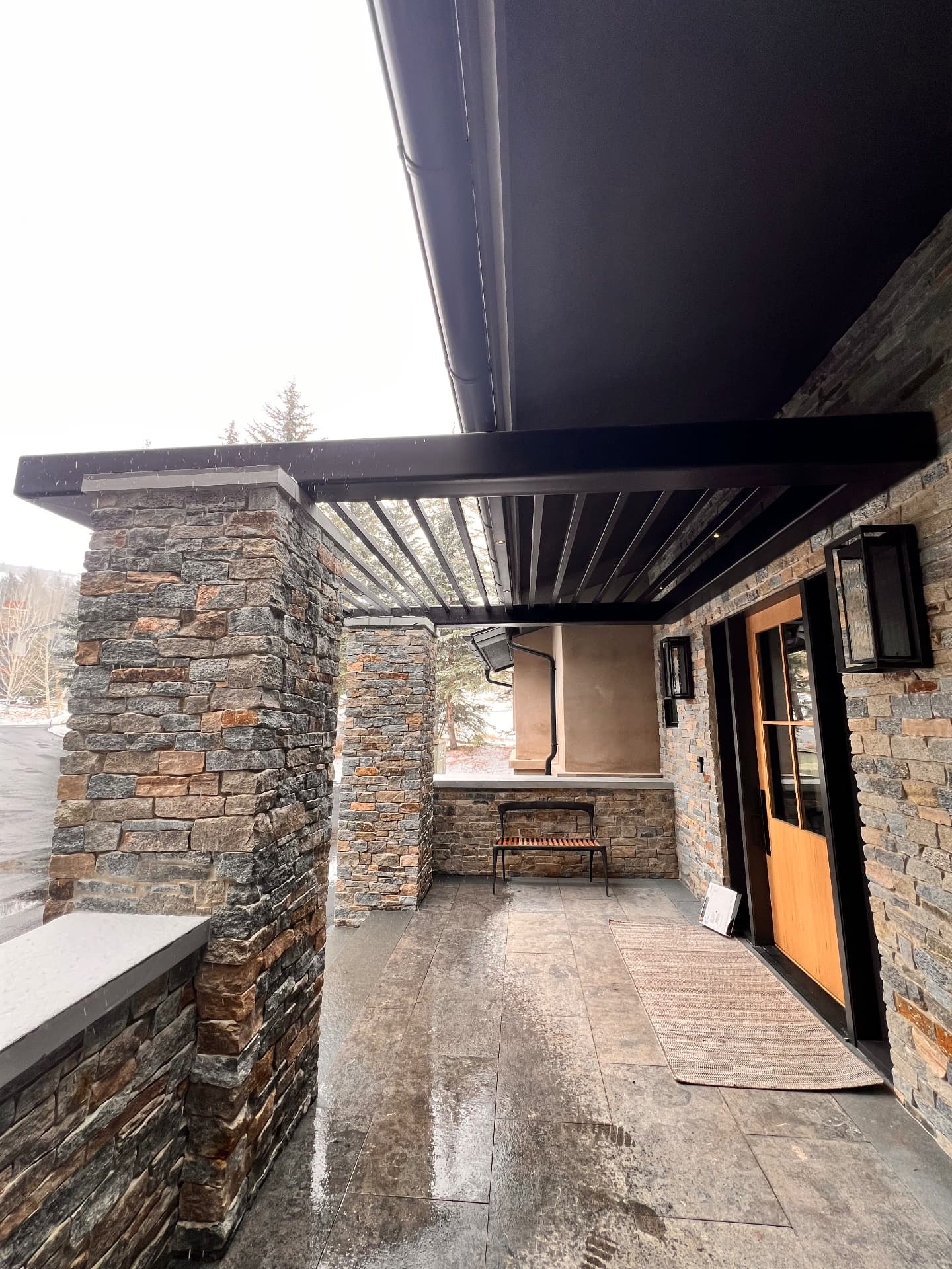 Black steel pergola attached to stone columns covering a covered stone patio entry with a wooden door