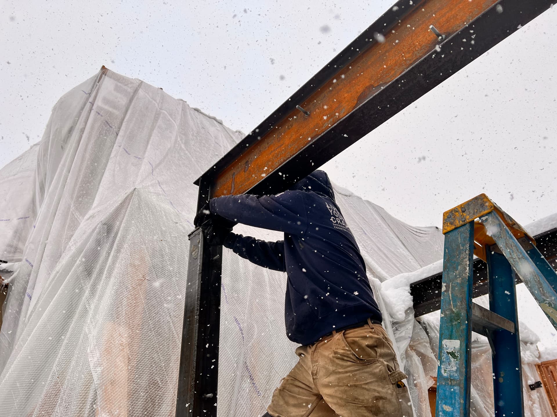 Welder guiding a weathered steel I-beam onto a support column during a winter structural install