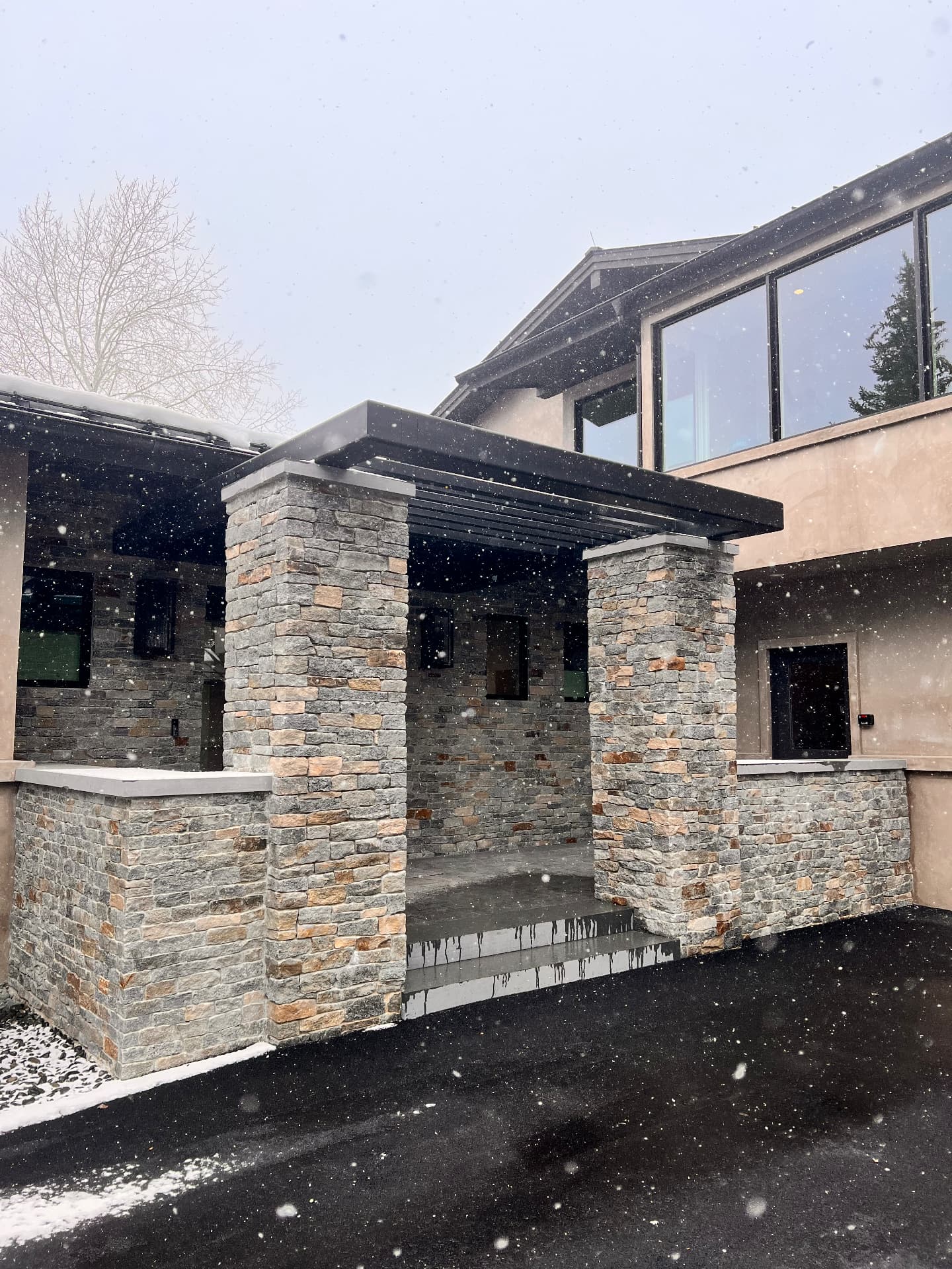 Custom black steel pergola and entry canopy supported by stone columns during snowfall